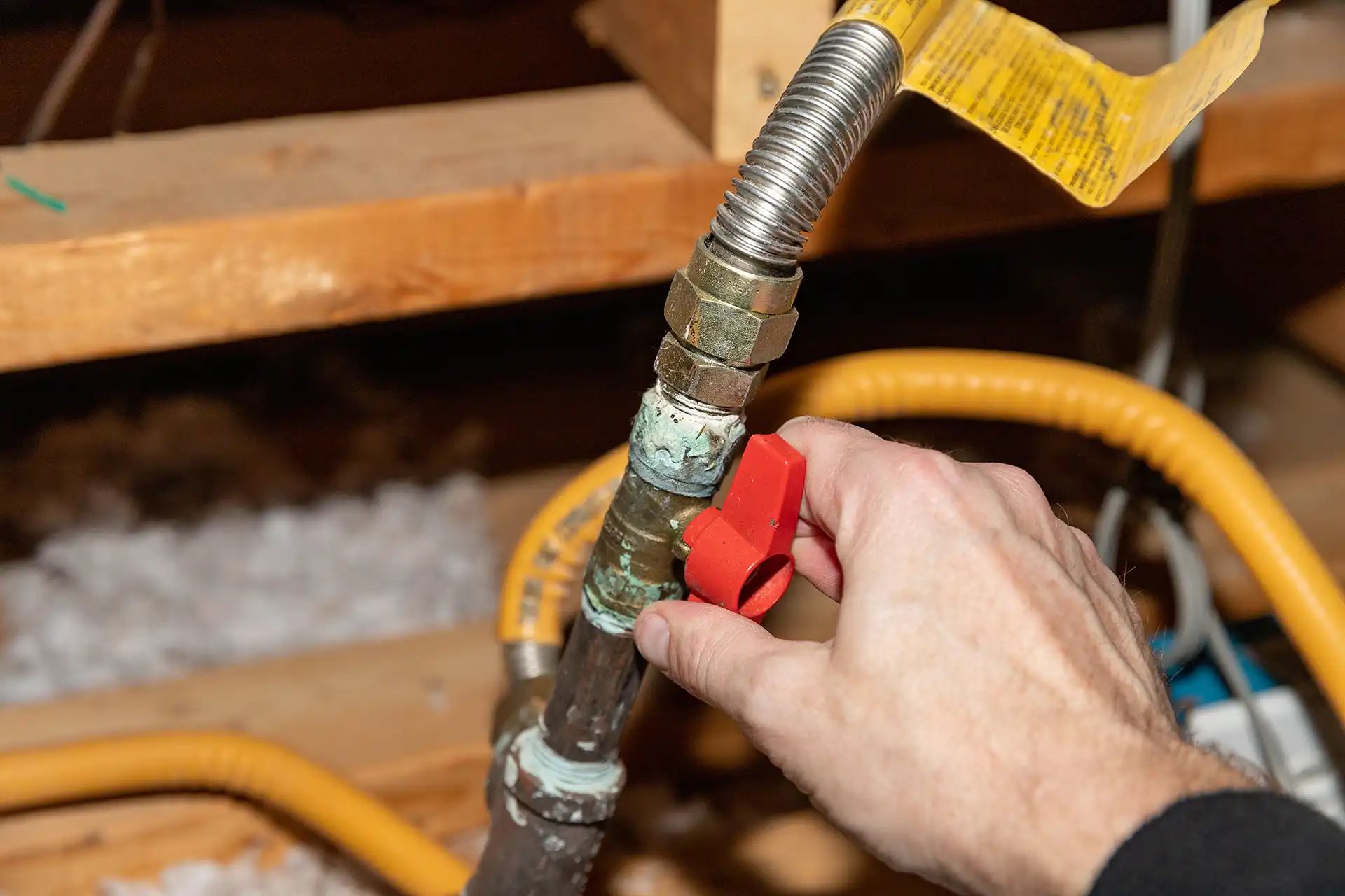 Technician's hand turning a red gas shutoff valve on a flexible gas line in an attic