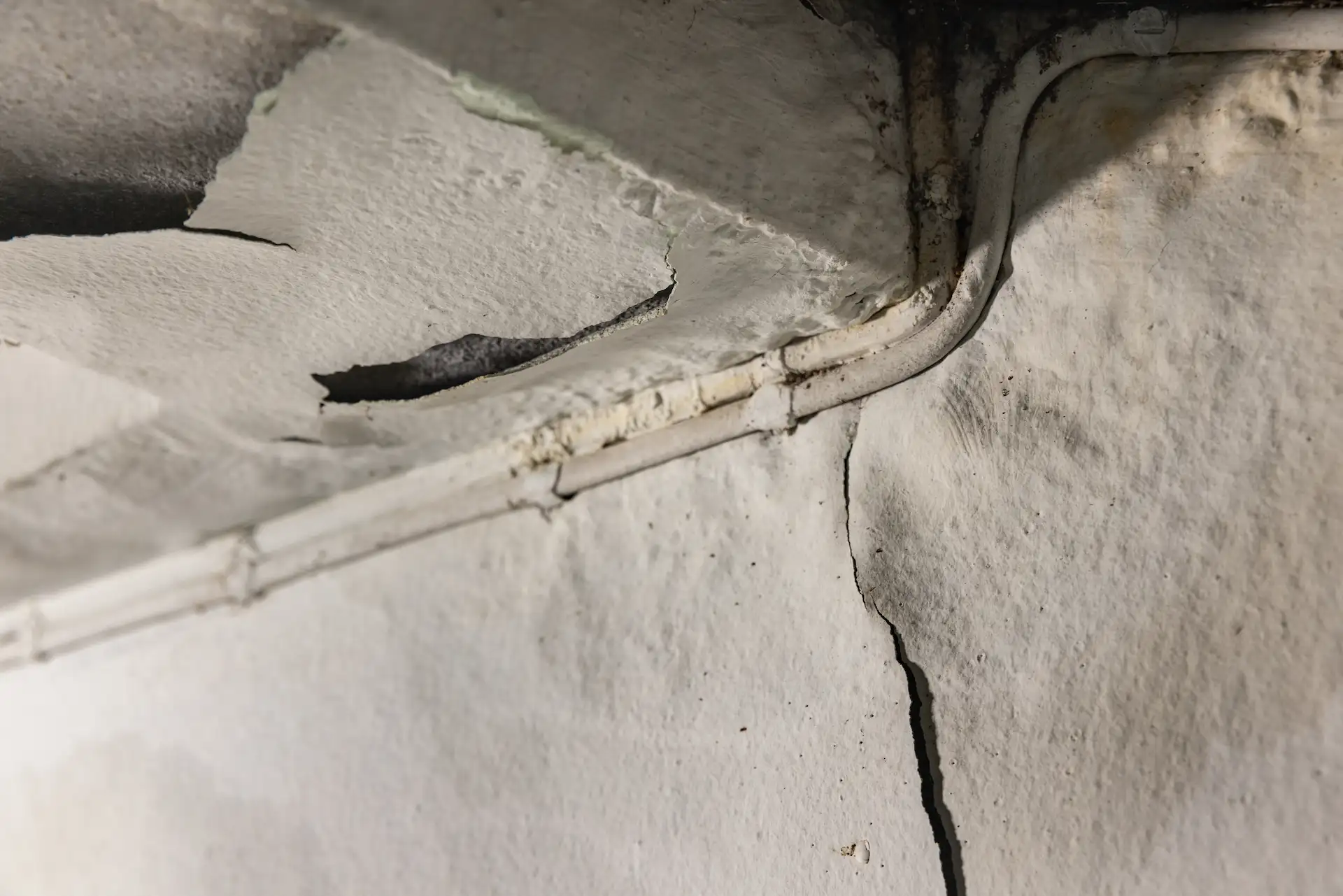 Close-up of a cracked foundation wall corner with a plumbing pipe running along the ceiling, showing water staining and cracking.