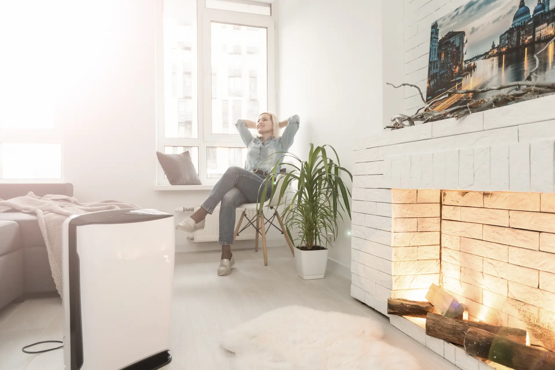 Woman relaxing in a chair by a sunny window near a fireplace, with a white air purifier running on the floor beside her.