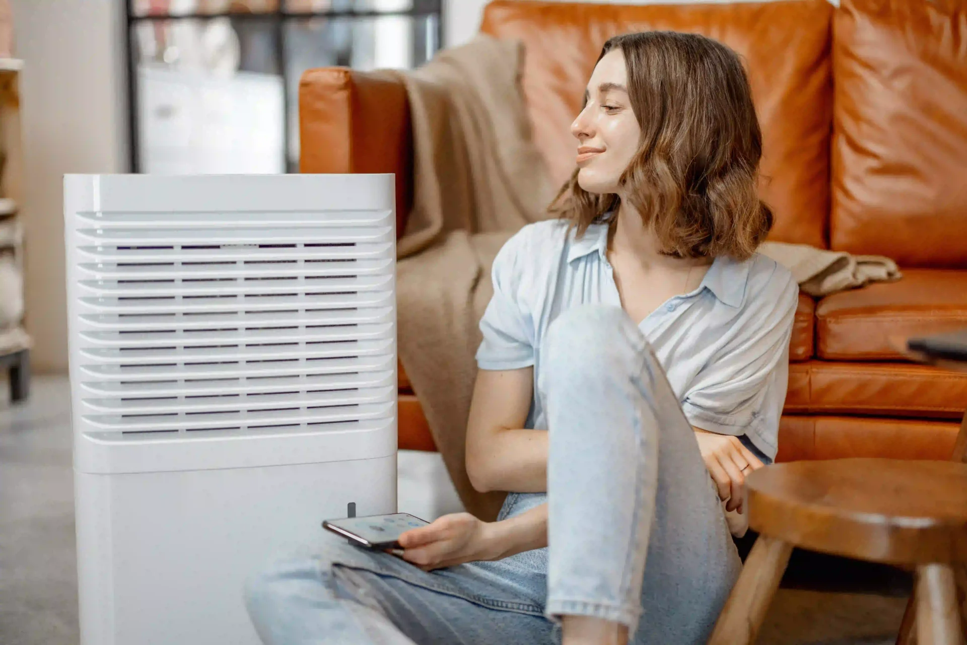 Woman relaxing on the floor near a leather couch beside a white air purifier, checking its settings on her phone.