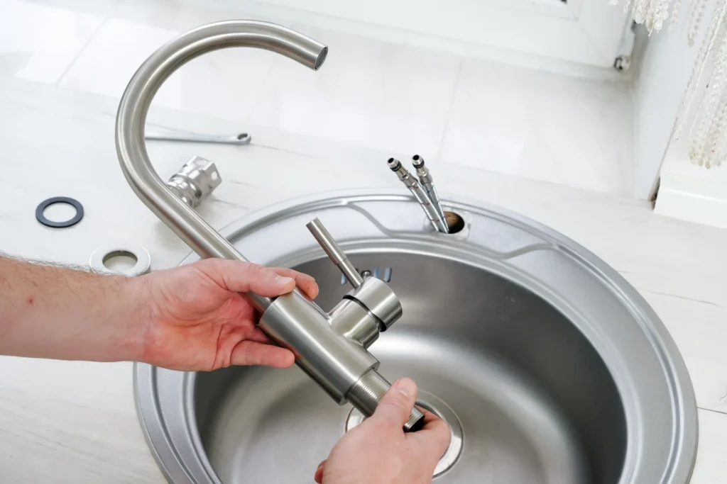 Plumber's hands installing a stainless steel kitchen faucet into a sink basin, with a wrench and supply lines nearby.
