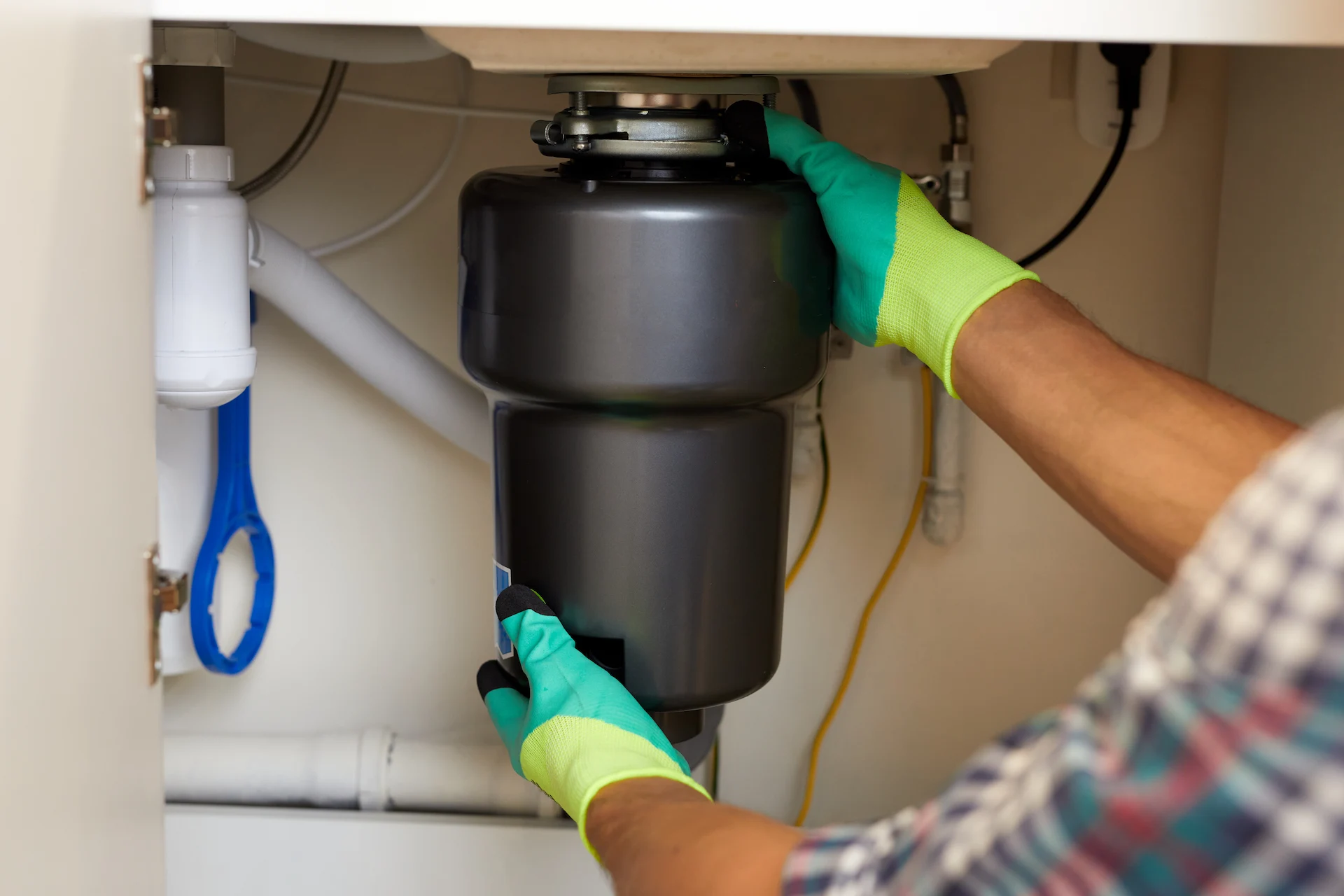 Gloved hands wearing green and yellow work gloves installing a black garbage disposal unit beneath a kitchen sink.