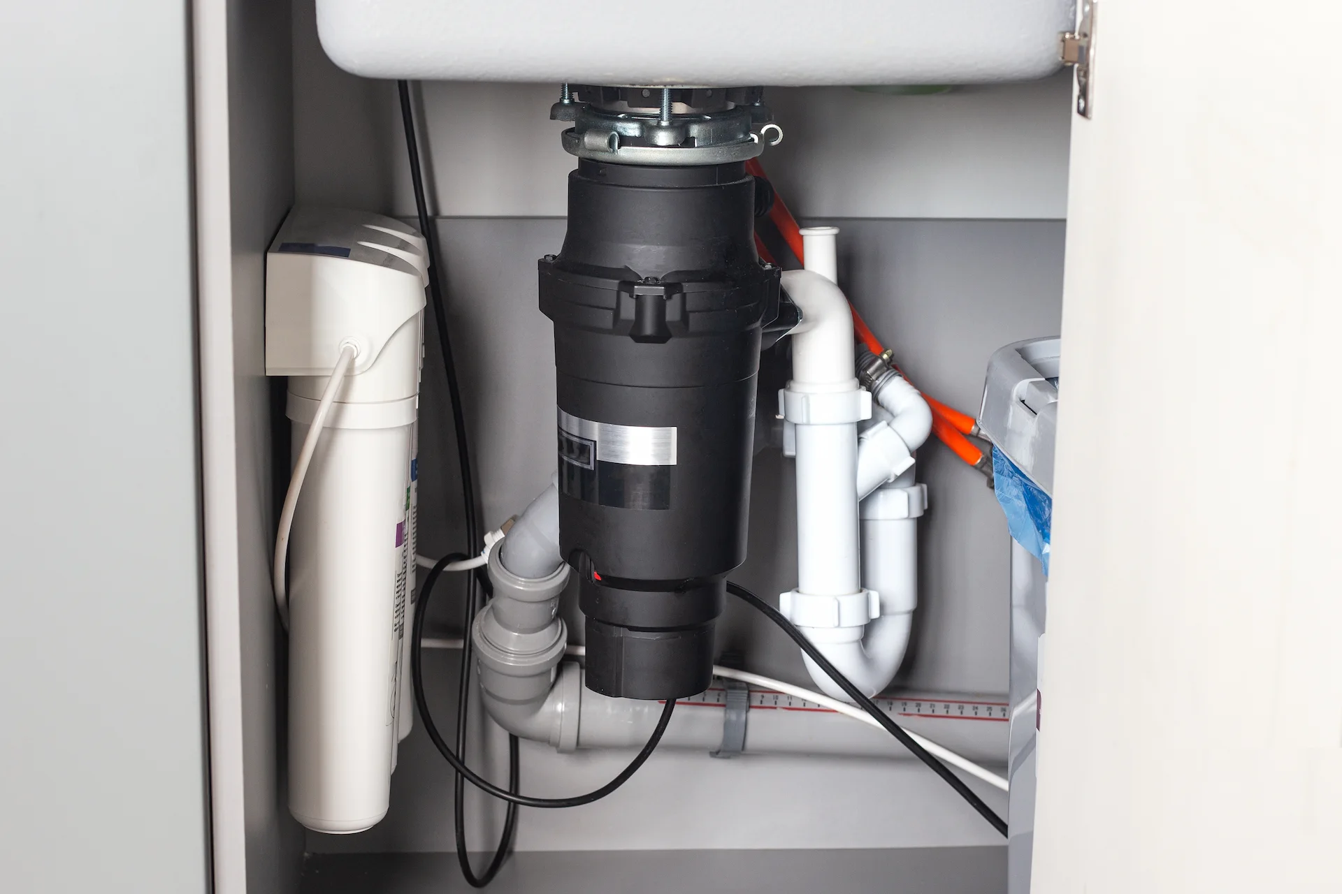 Garbage disposal unit installed under a kitchen sink beside a water filtration cartridge, with white PVC drain pipes nearby.