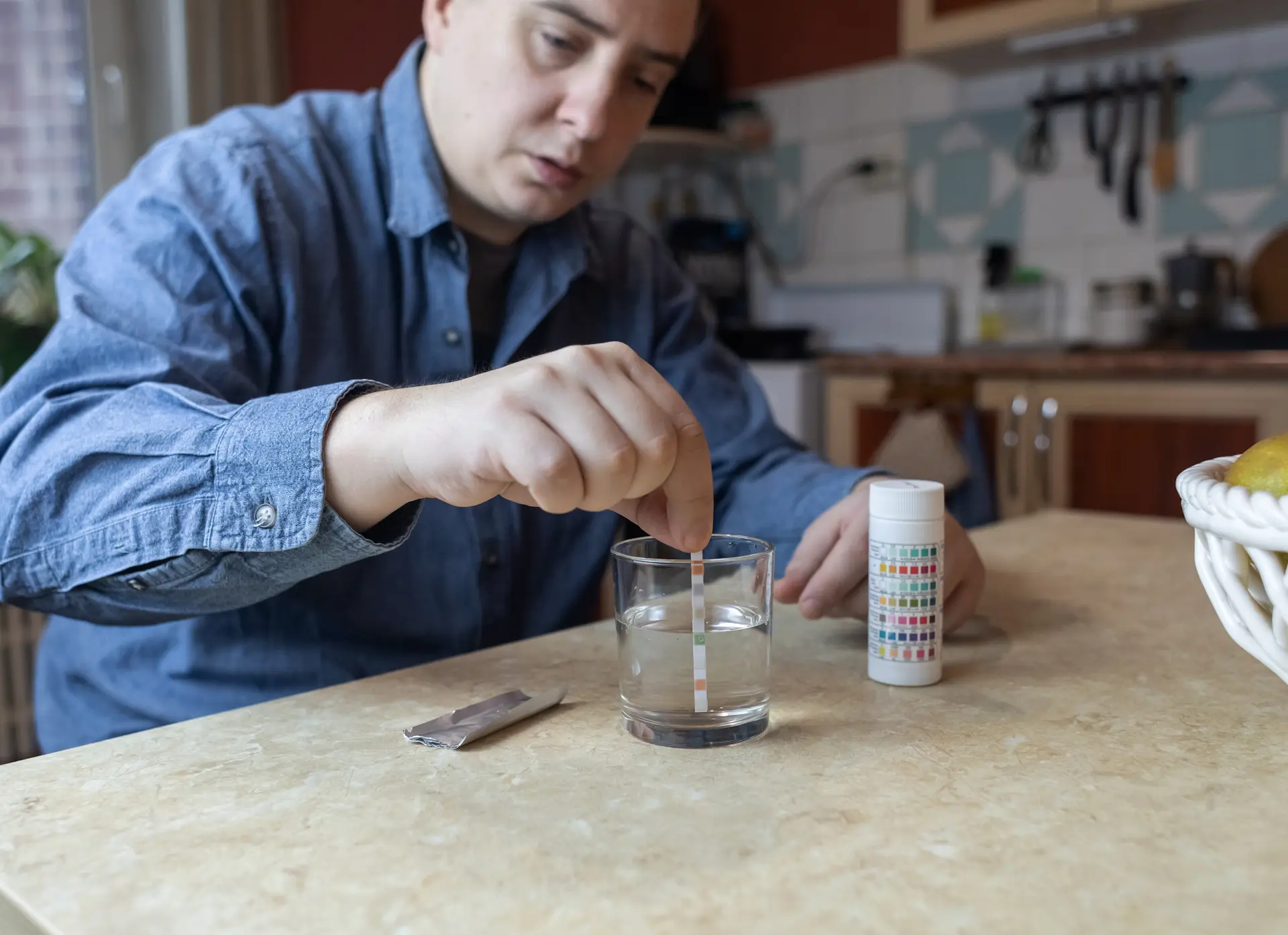 Man testing water quality with a color-coded test strip dipped in a glass of water on a kitchen counter.
