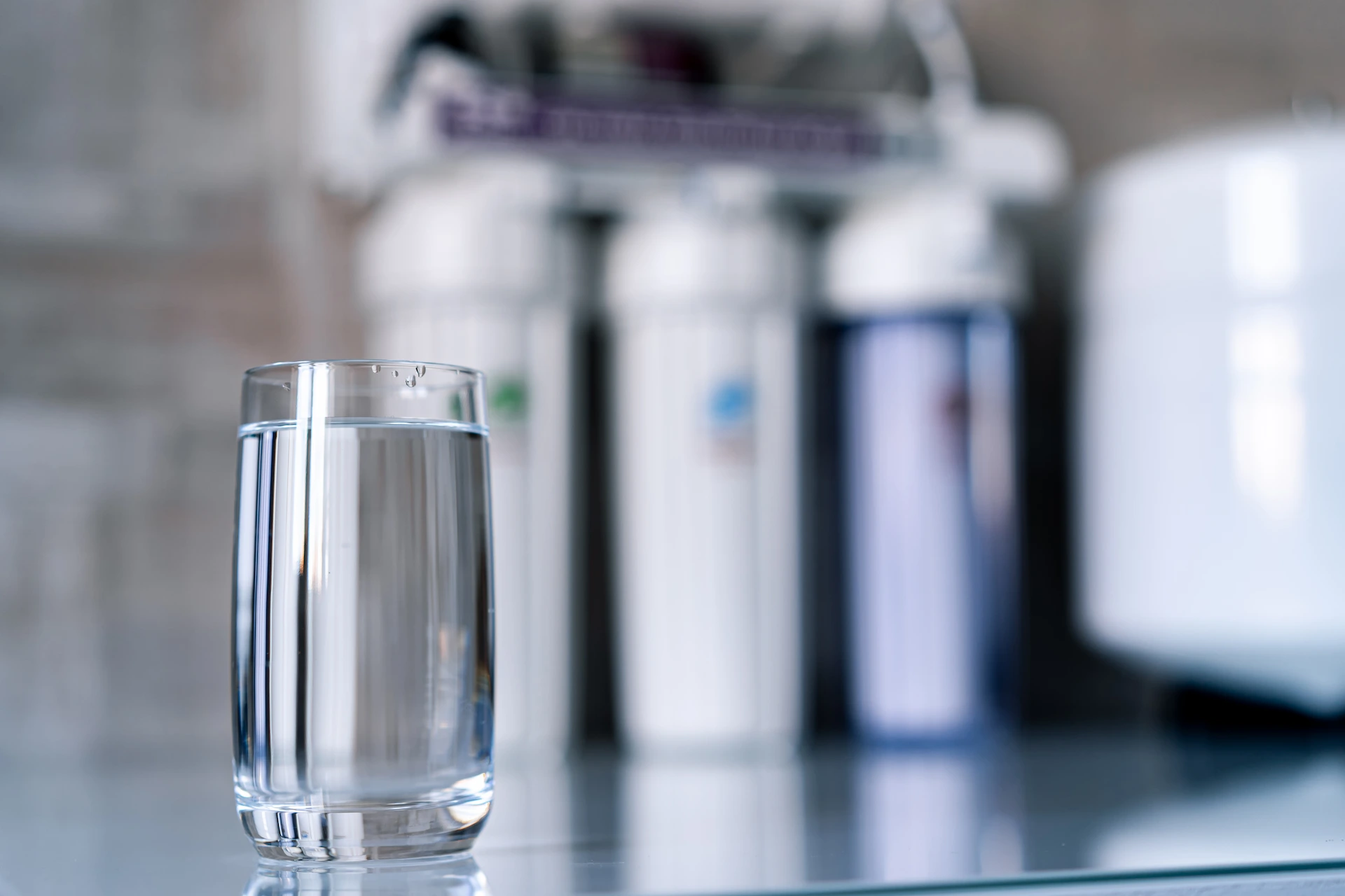 Glass of clear water on a counter in front of a blurred whole-house water filtration system.